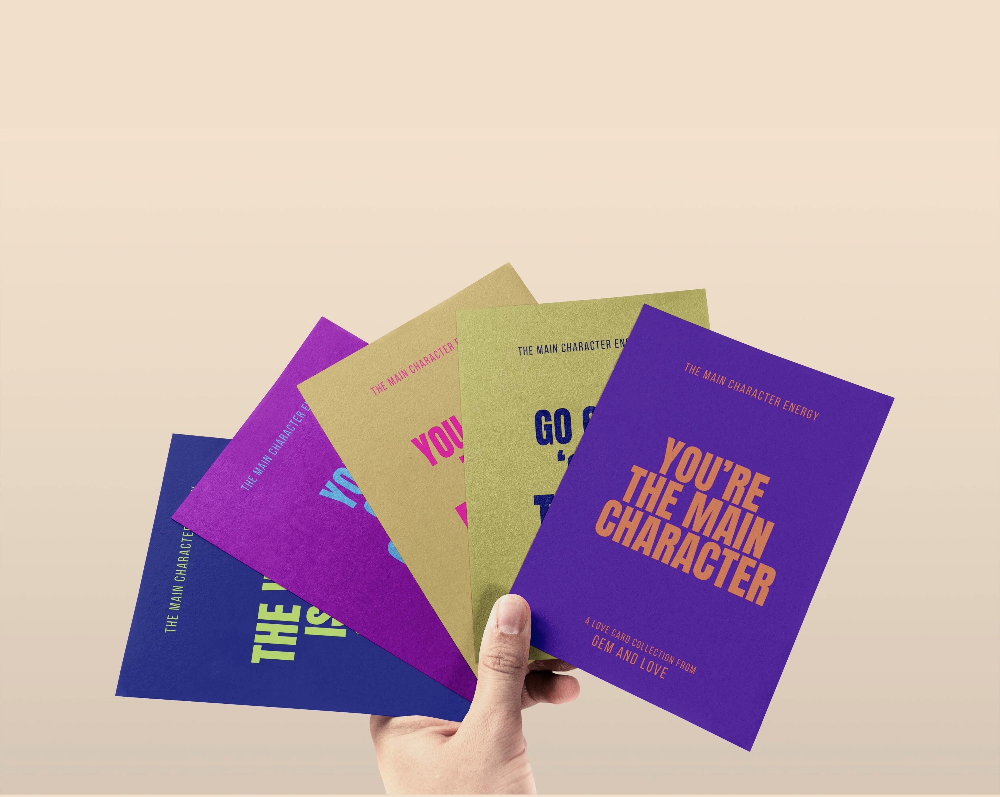 Hand holding a purple card with 'You're the Main Character' text against a beige background